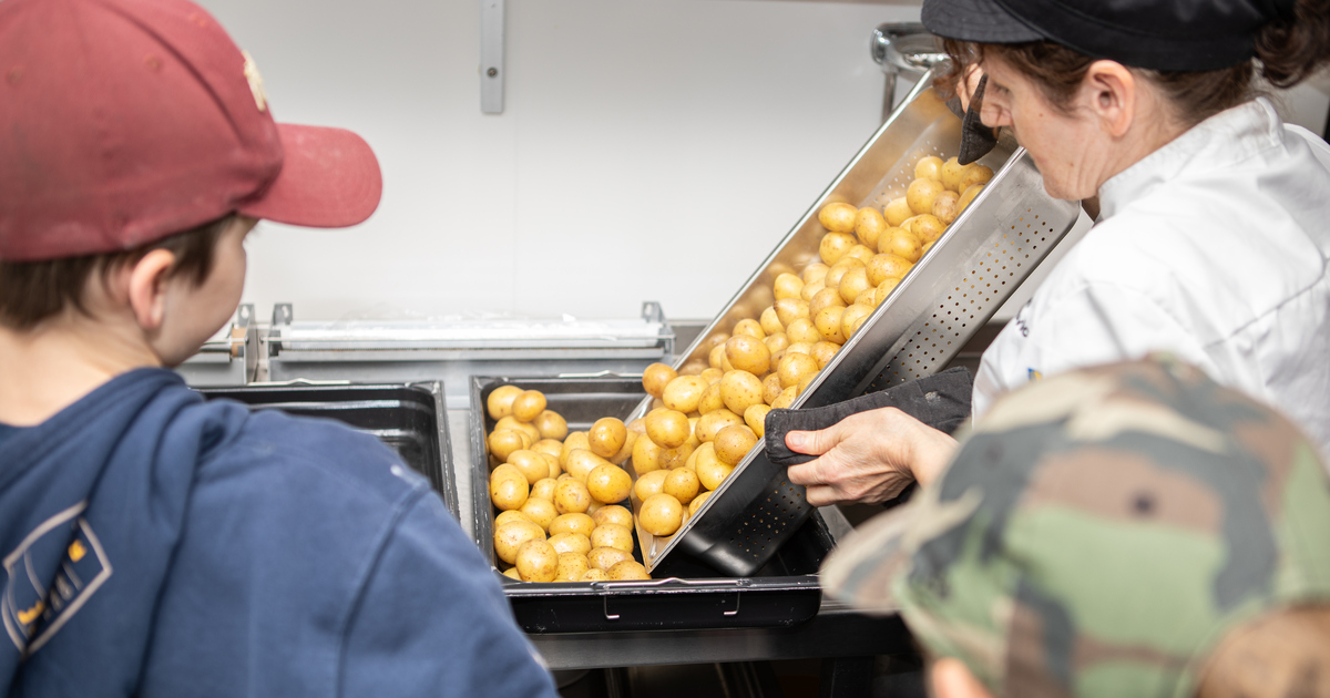 En kock häller potatis från en behållare till en annan medan två skolbarn tittar på.