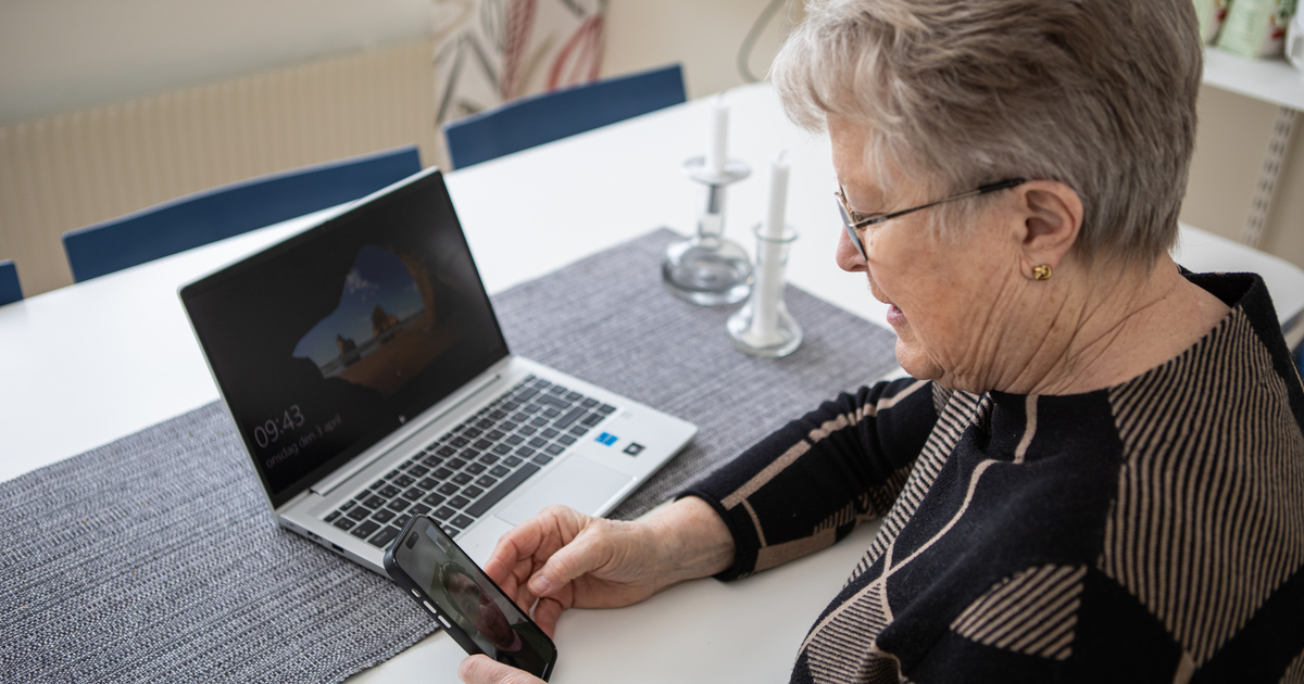 En äldre kvinna sitter framför en dator med en telefon i handen. På telefonens skärm syns det att hon är mitt i ett videosamtal.